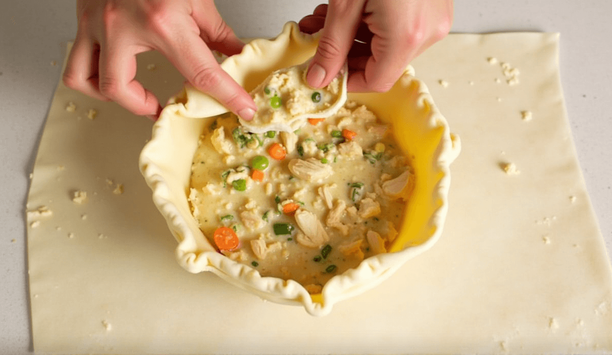 A person assembling a chicken pot pie: spreading chicken and vegetable mixture into a pie crust, with a second crust being placed over the top, ready to be crimped.